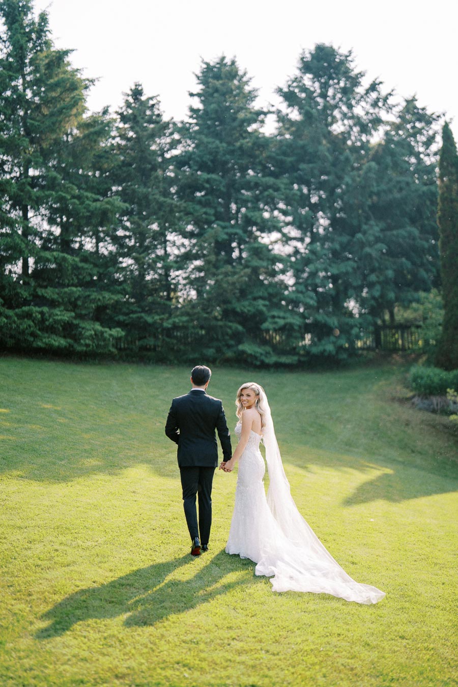 Bride and groom holding hands, walking across a sunlit, grassy field with evergreen trees in the background, showcasing a serene outdoor wedding setting.