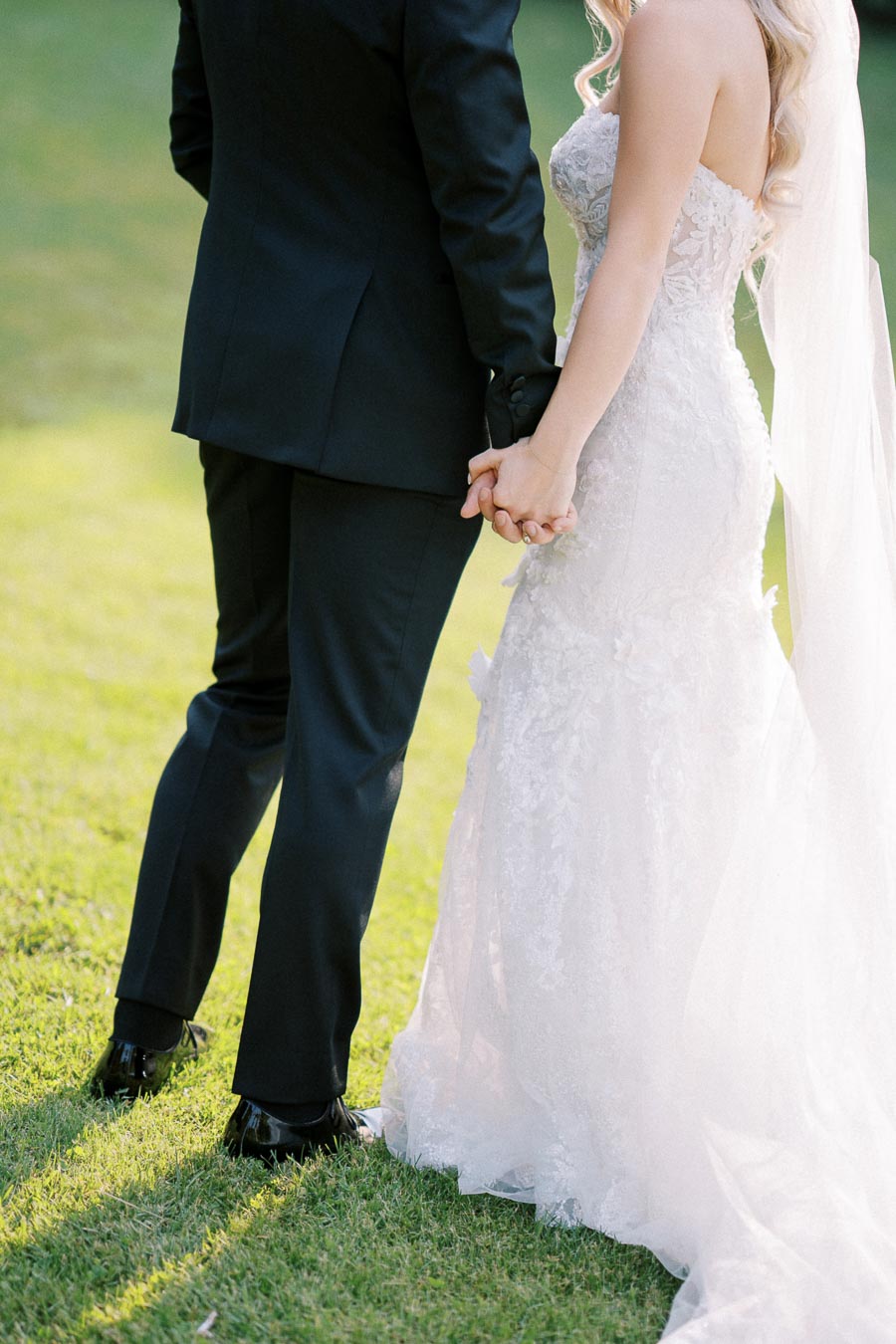 Elegant bride in lace gown holding hands with groom in black suit on a sunny day.