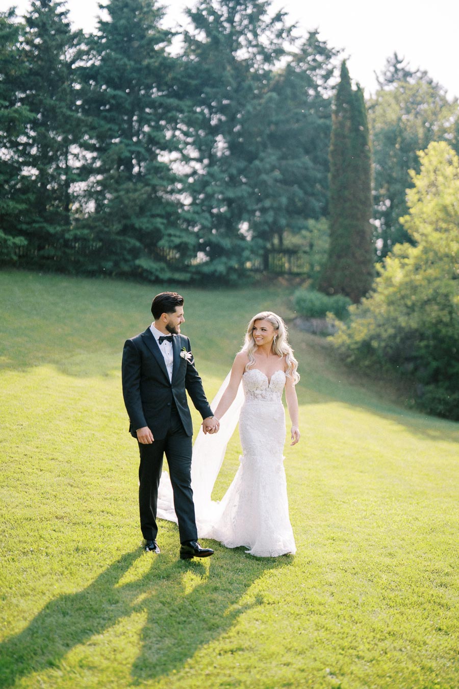 Bride and groom walking hand-in-hand on a sunny day, surrounded by lush green trees and grass, outdoor wedding photography.