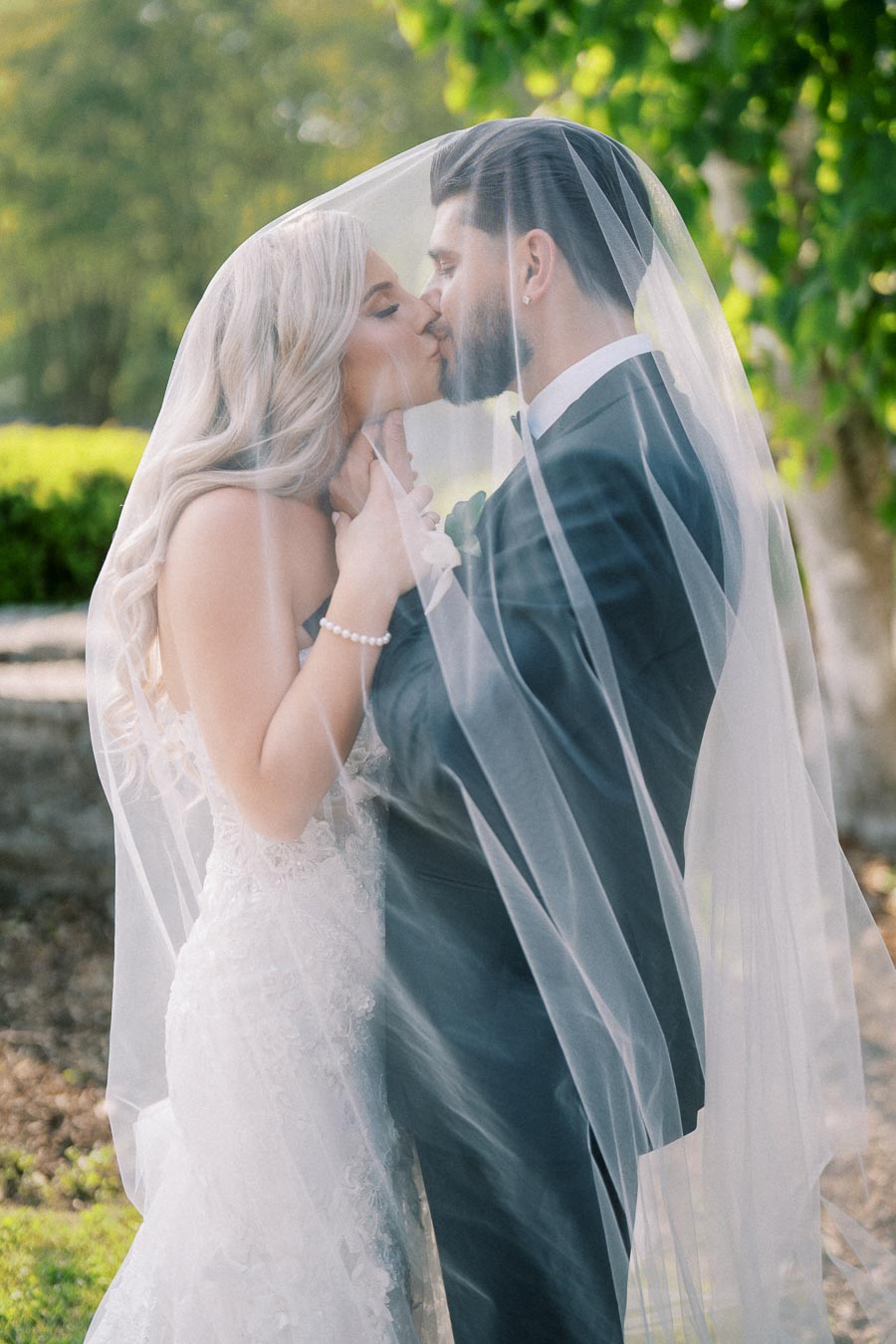A bride and groom share a romantic kiss under a veil in a garden setting, highlighting a beautiful wedding moment with the bride in a lace dress and the groom in a black suit.