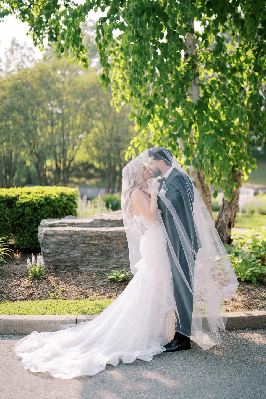 A bride and groom share a romantic kiss under a sheer veil during an outdoor wedding ceremony. The bride wears an elegant white gown while the groom is dressed in a formal suit. They are surrounded by lush greenery and natural sunlight, creating a picturesque wedding setting.