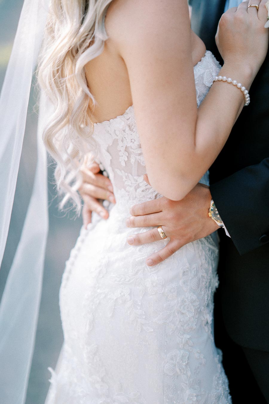 Close-up of a bride and groom embracing, showcasing the intricate lace details of the bride's wedding dress and the groom's hand adorned with a wedding ring, symbolizing love and commitment on their special day.
