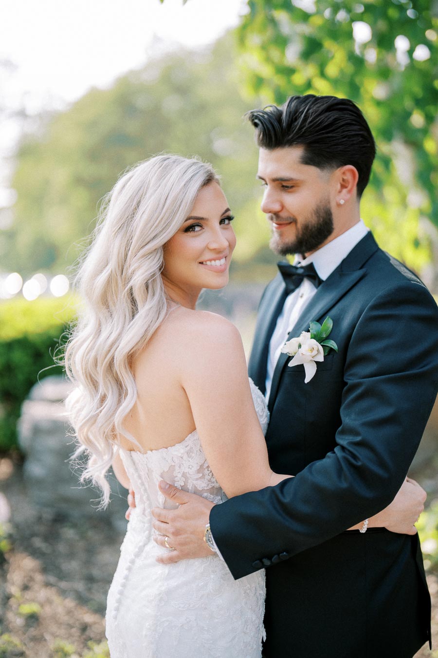 A bride with long, blonde curls smiles at the camera while embracing the groom in a black tuxedo with a boutonniere, set against a lush, green outdoor backdrop.