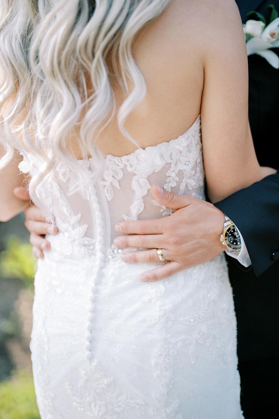 Close-up of a bride in an elegant lace wedding dress with long, wavy blonde hair, embraced by a groom wearing a black suit and a gold watch, showcasing their wedding rings.