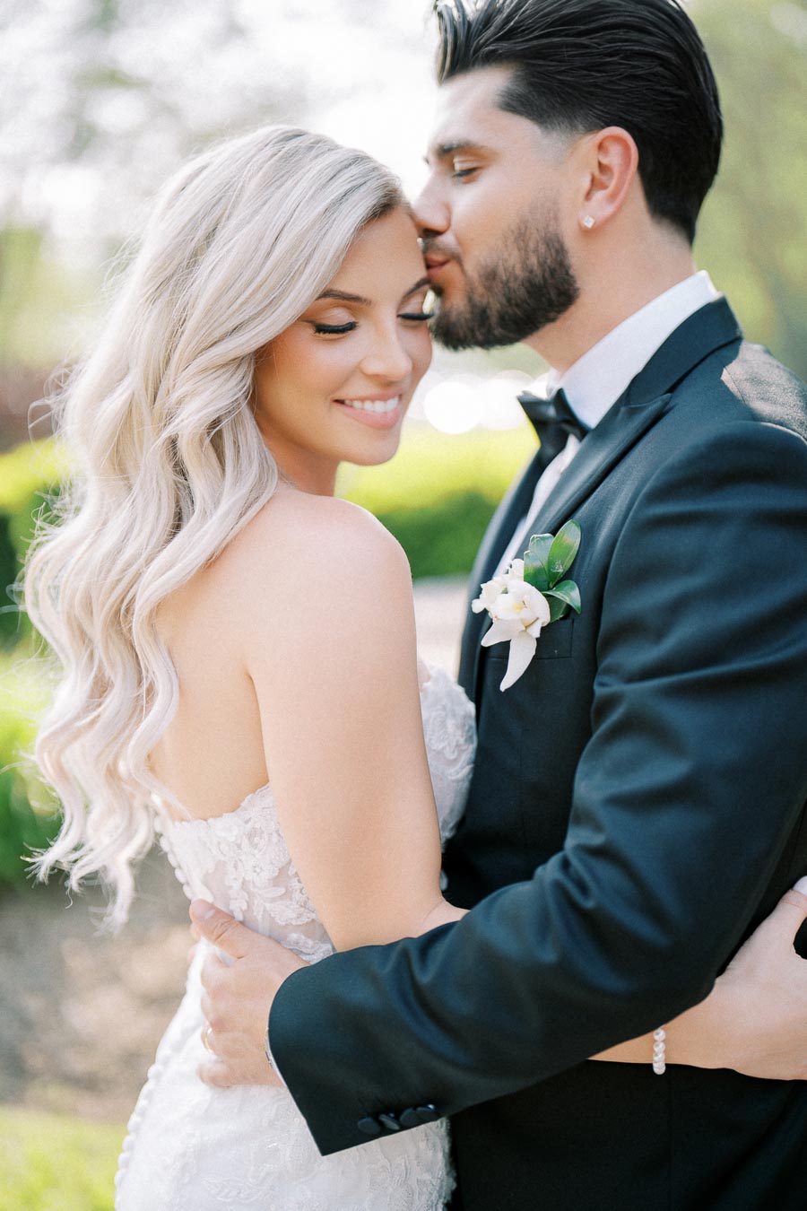 A romantic wedding moment capturing a groom in a black tuxedo kissing the bride with long blonde hair in a white lace dress, set against a lush outdoor backdrop.