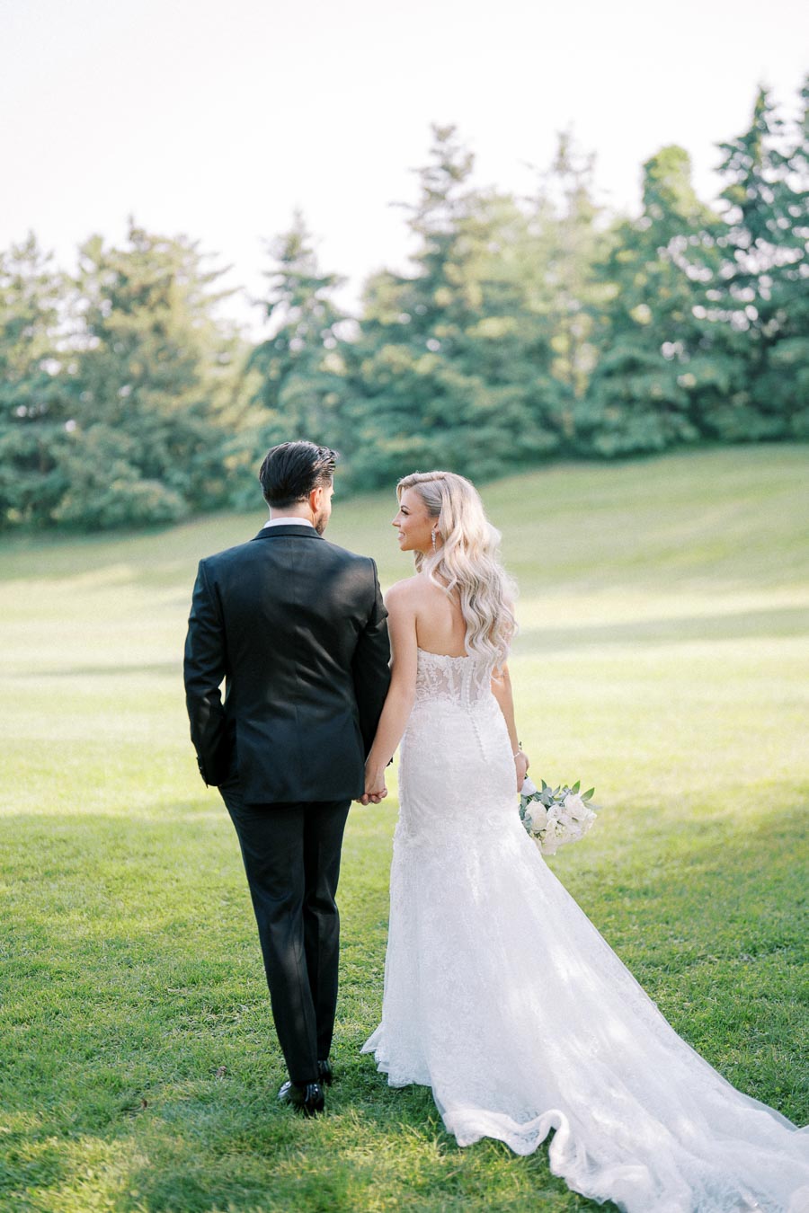 Bride and groom walking hand in hand on a grassy field, with the bride wearing a white lace wedding gown, and the groom in a black suit, surrounded by a serene landscape with trees in the background.