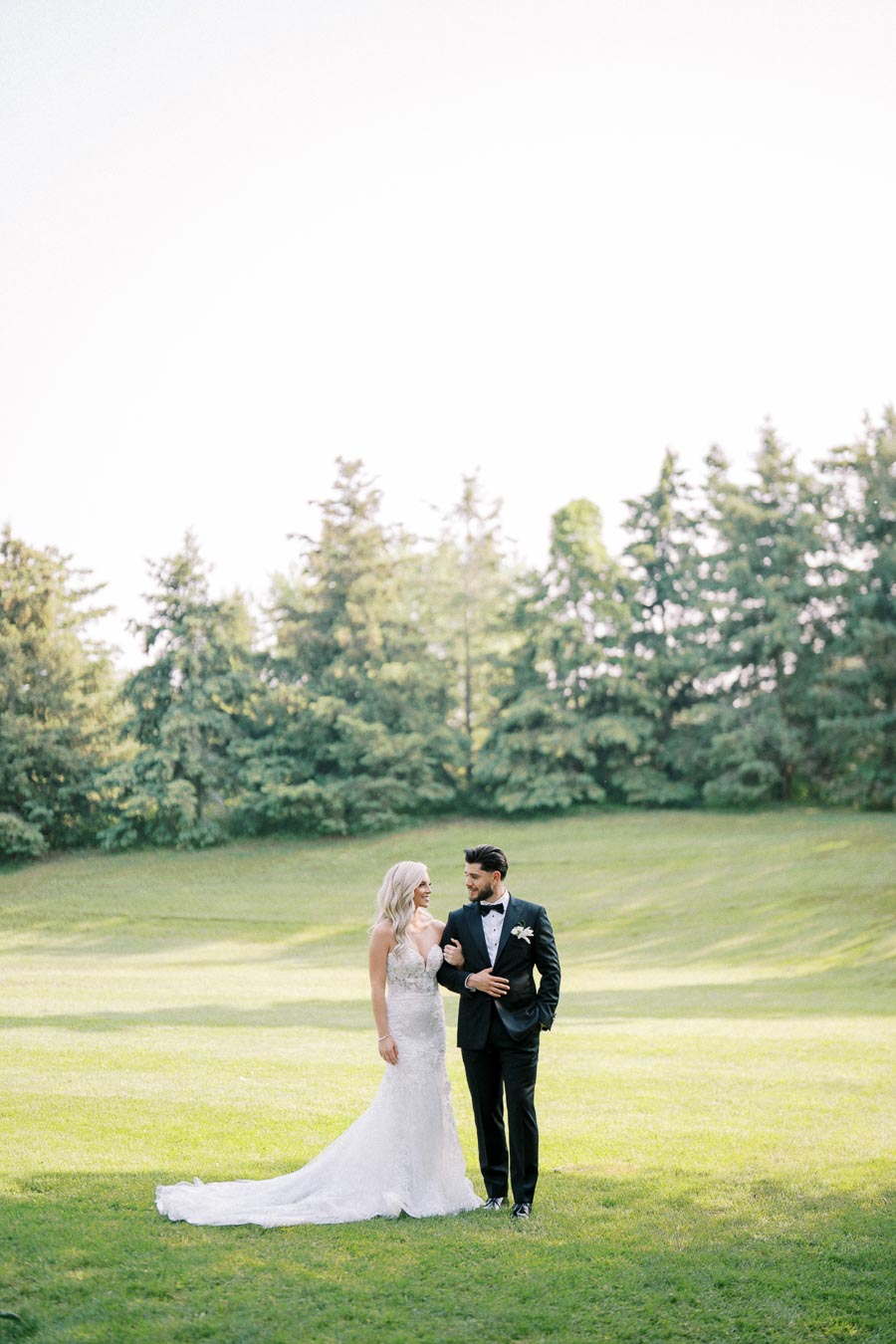 A bride in a white lace gown and groom in a black tuxedo walk hand in hand across a lush, green field with tall trees in the background, under a bright, clear sky.