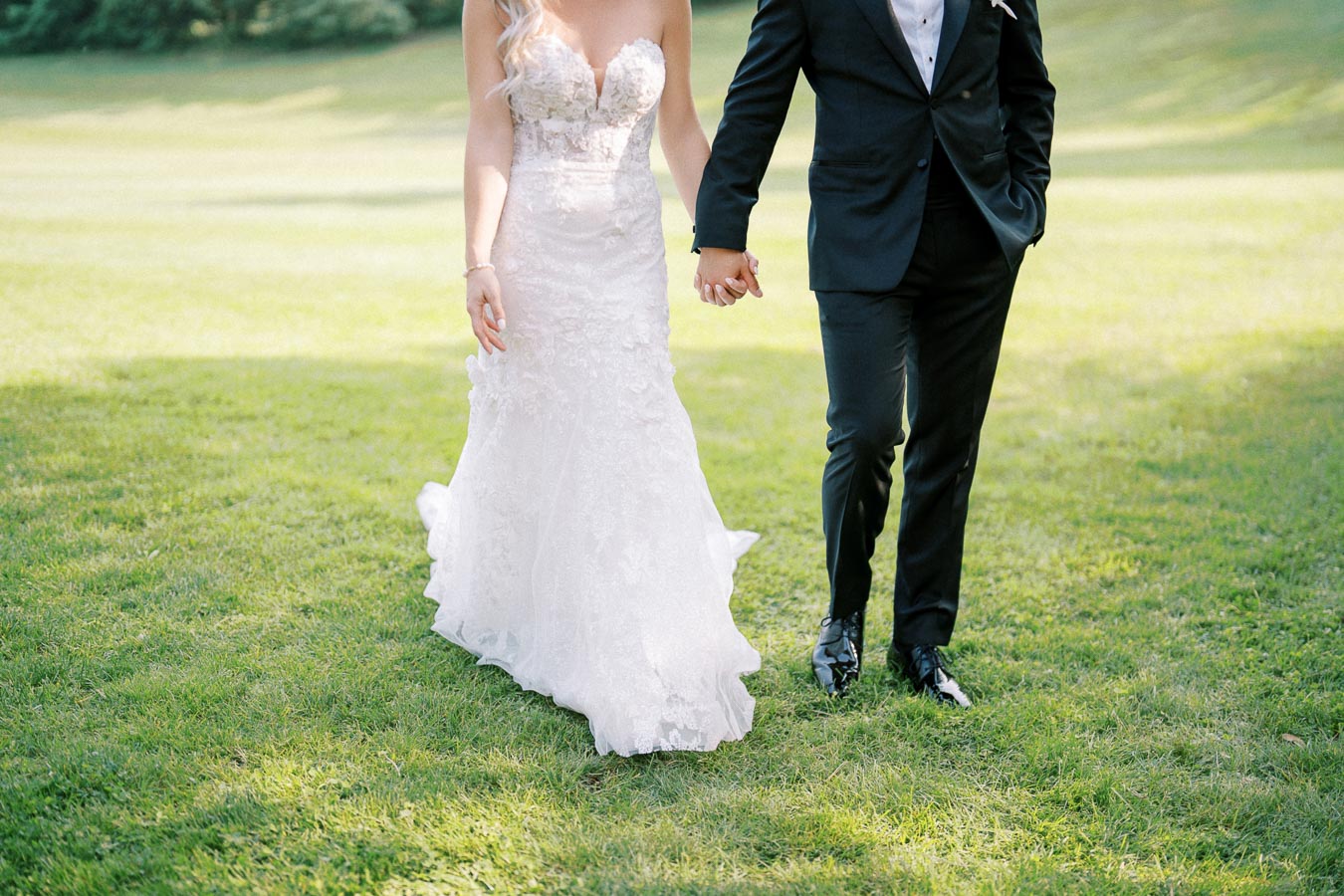 A bride in a lace wedding dress and a groom in a black suit holding hands while walking on green grass outdoors.