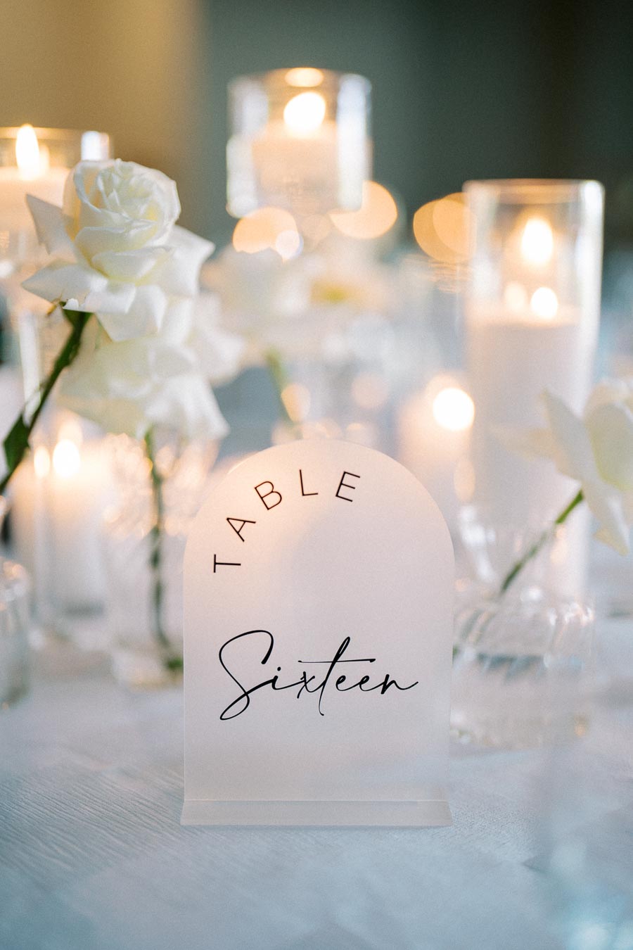 Elegant table setting with candles and white roses, featuring a frosted table number sign reading Table Sixteen. Perfect for a wedding or formal event ambiance.