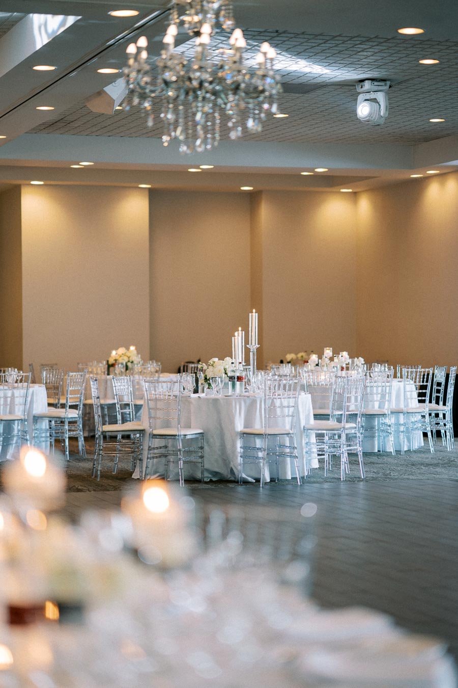 Elegant wedding reception hall with white table linens, crystal chandeliers, and chic transparent chairs arranged around round tables, adorned with tall candle holders and floral centerpieces.