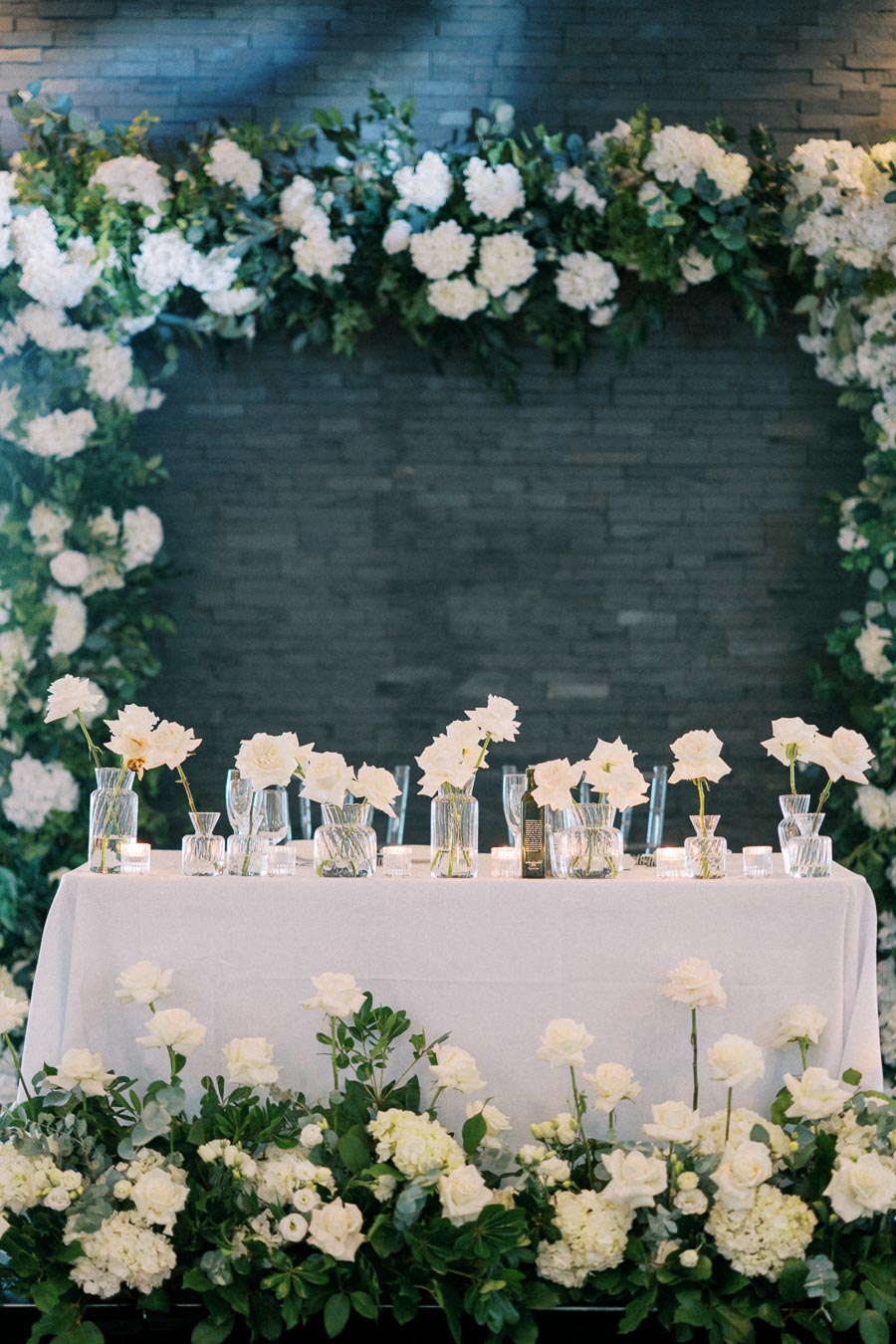 Elegant wedding reception table decorated with white roses and glass vases, surrounded by lush greenery and floral arrangements, set against a dark brick backdrop.