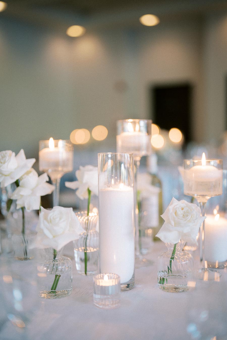 Elegant wedding table centerpiece with white roses in glass vases and tall, lit candles, creating a romantic ambiance.