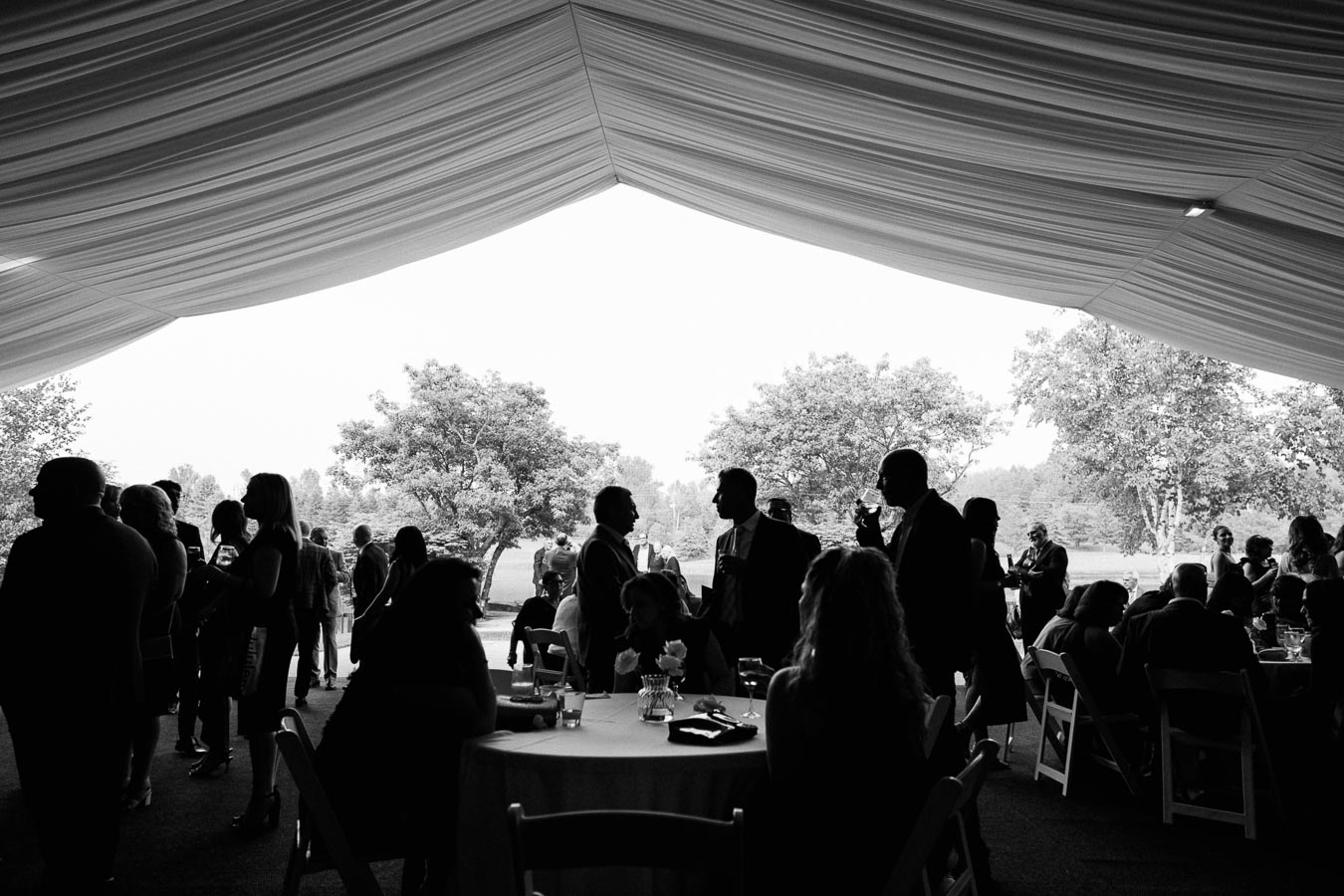 Black and white image of people socializing at an outdoor event under a tent, with trees in the background.