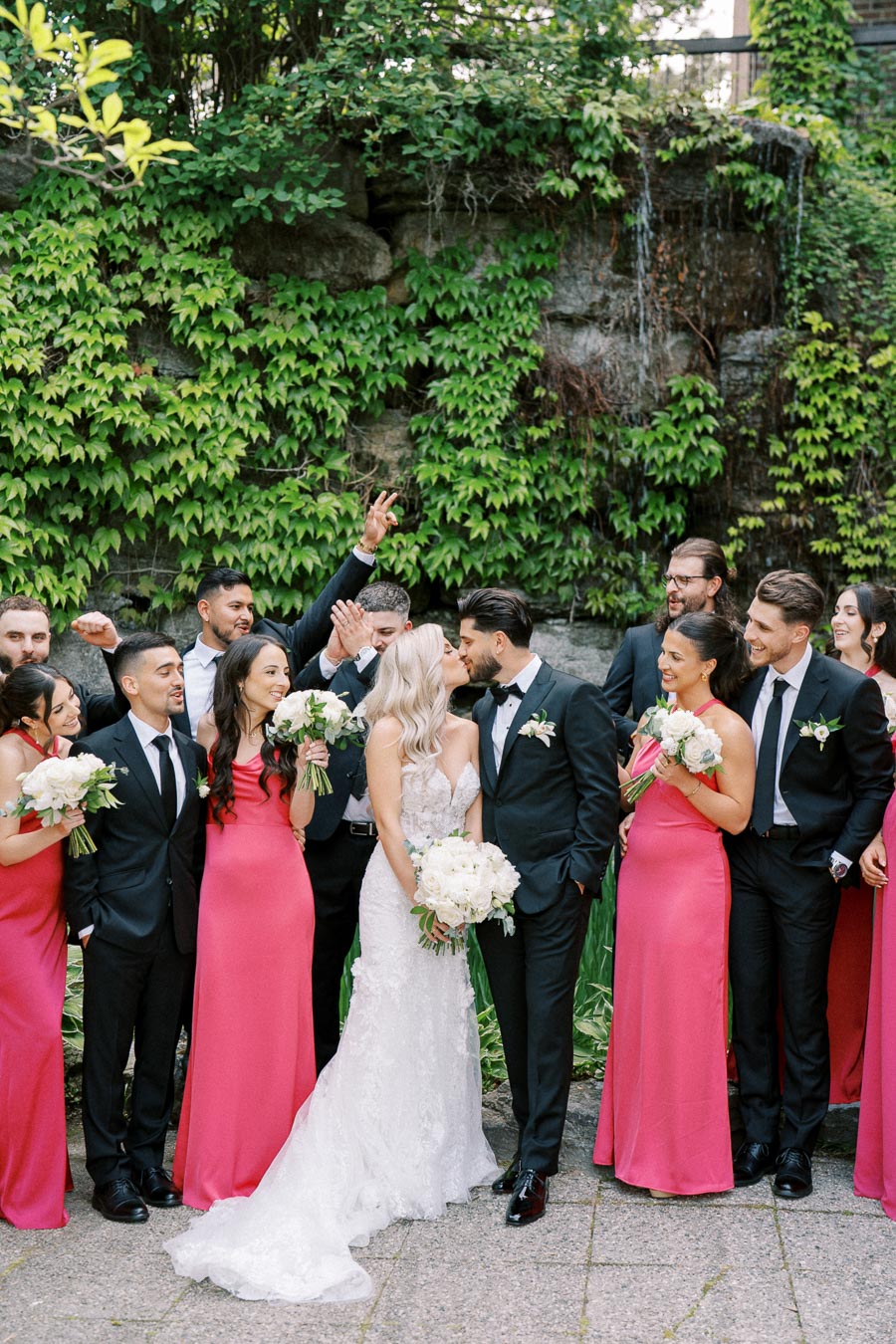 A joyful wedding party poses outdoors, featuring a bride in a white lace gown and a groom in a black tuxedo kissing, surrounded by bridesmaids in pink dresses and groomsmen in black suits holding bouquets, against a lush green ivy backdrop.