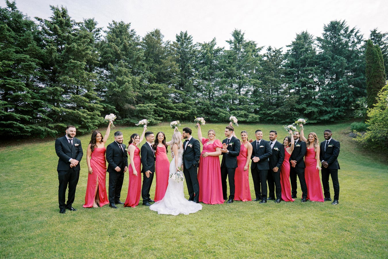 A joyful wedding party posing outdoors, featuring bridesmaids in coral dresses and groomsmen in black suits, with lush greenery and trees in the background.