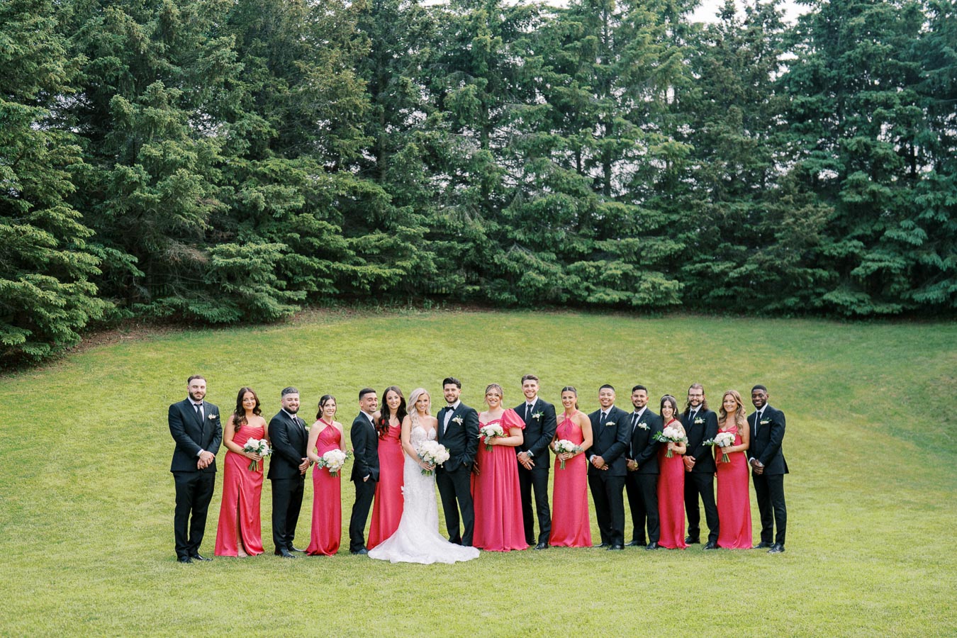 Wedding party posed on a lush green lawn with bridesmaids in red dresses and groomsmen in black suits, standing in front of a backdrop of tall evergreen trees, capturing a joyful outdoor celebration.