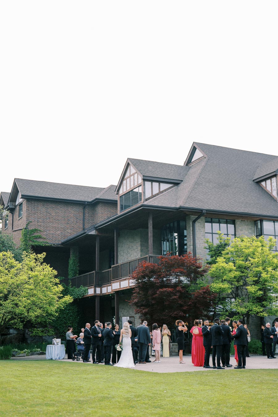 A wedding reception outdoors at a large brick estate, with guests in formal attire gathered on the lawn, surrounded by lush greenery and trees.