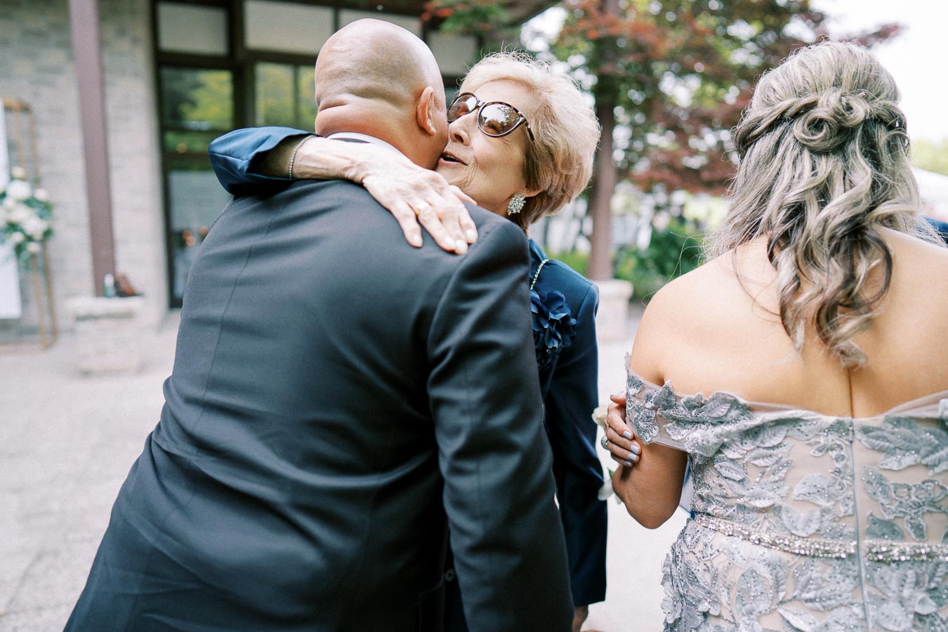 Elderly woman in sunglasses warmly embraces a bald man in a suit, with a woman in a detailed lace dress standing beside them, outdoors.