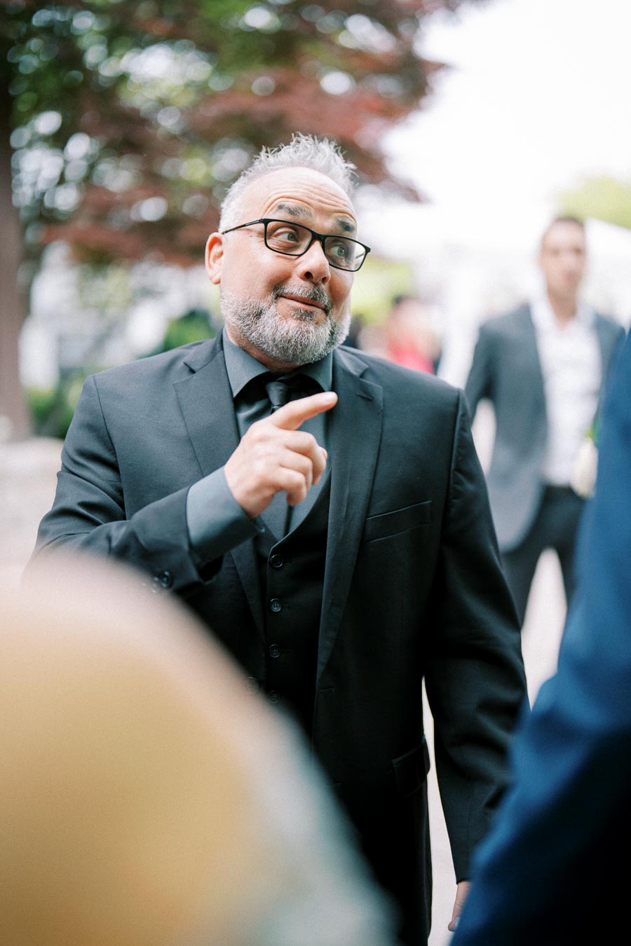 A man with glasses in a suit making an expressive gesture outdoors, with a blurred background of trees and people in formal attire.