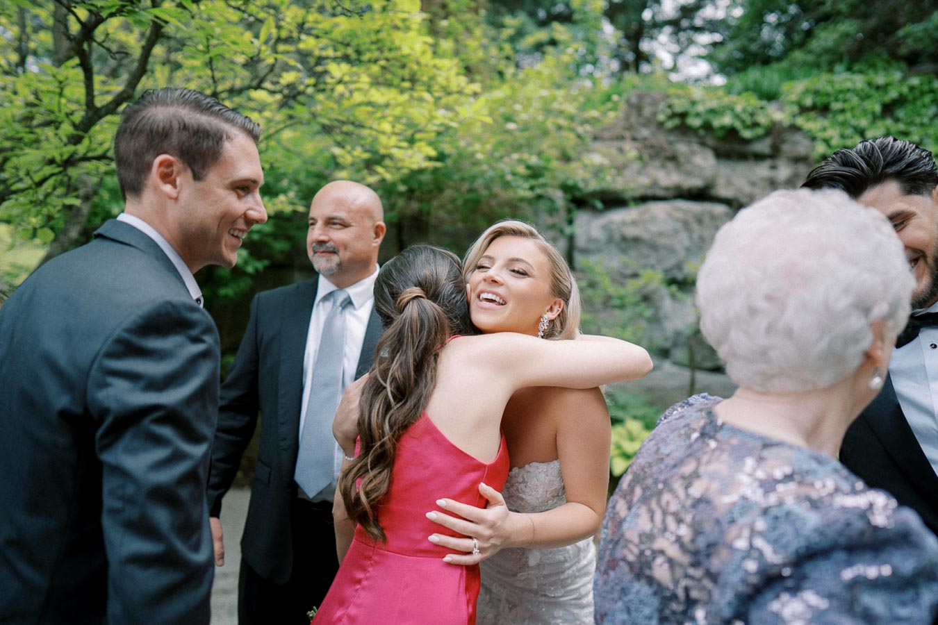 A joyful bride in a white wedding dress hugs a guest wearing a red dress outdoors, surrounded by smiling friends and family members dressed in formal attire, with a lush green garden setting in the background.