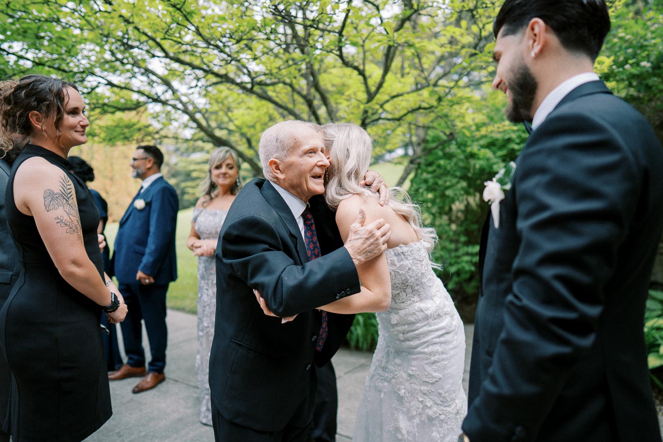 Wedding celebration with guests exchanging hugs and smiles outdoors in a green garden setting. A bride in a lace gown warmly embraces an older gentleman in a suit, surrounded by elegantly dressed attendees.