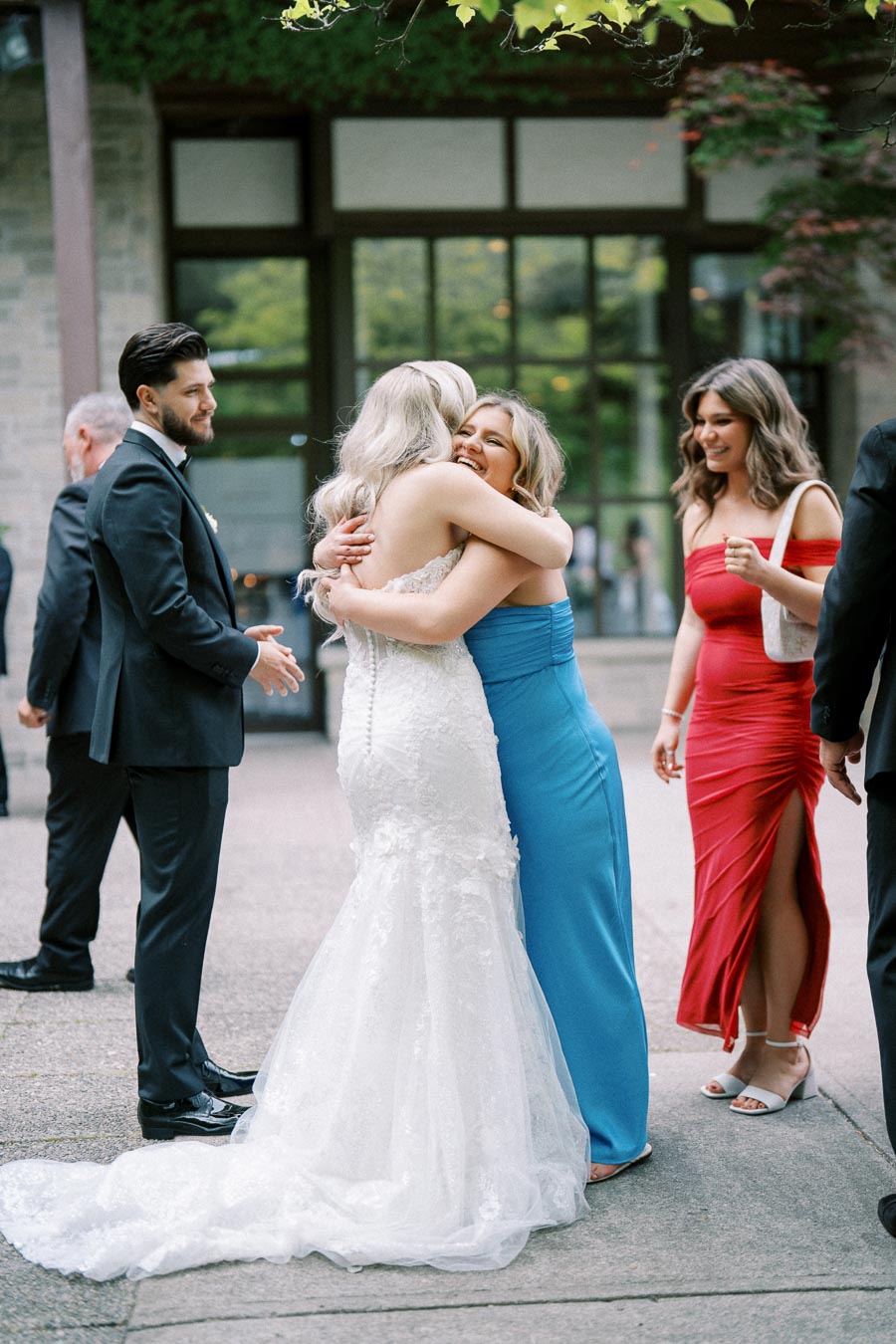 A bride in a white wedding dress embraces a smiling bridesmaid in a blue gown outdoors, with other elegantly dressed guests nearby, creating a joyful and celebratory wedding atmosphere.