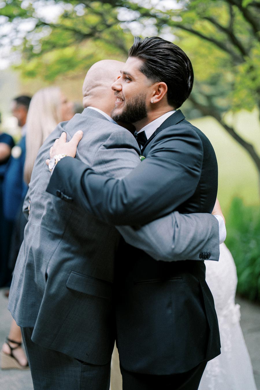 Two men in suits embracing warmly outdoors, symbolizing friendship and celebration at a wedding event with blurred nature in the background.