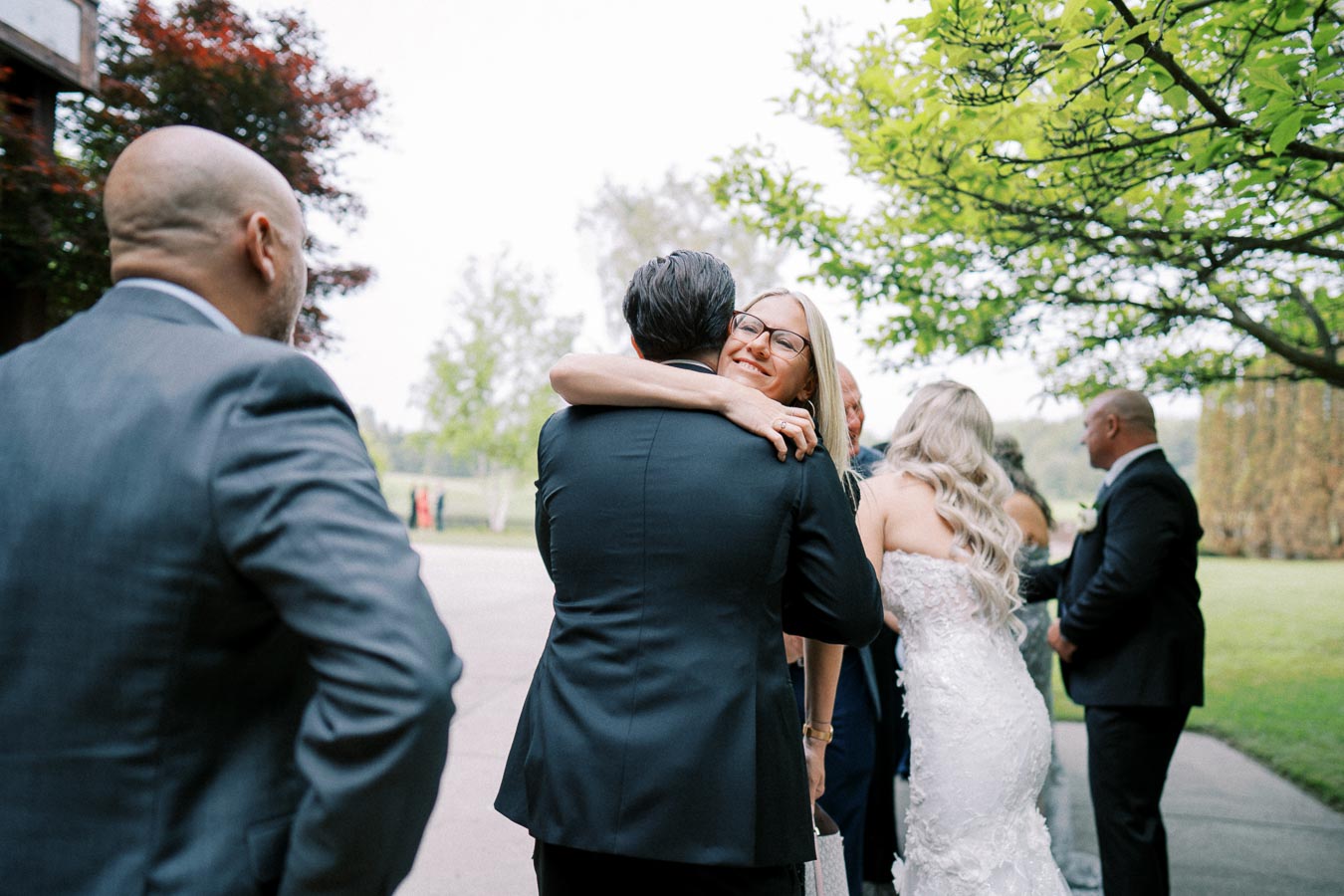 A joyful outdoor wedding scene with elegantly dressed guests greeting and embracing, surrounded by lush greenery.