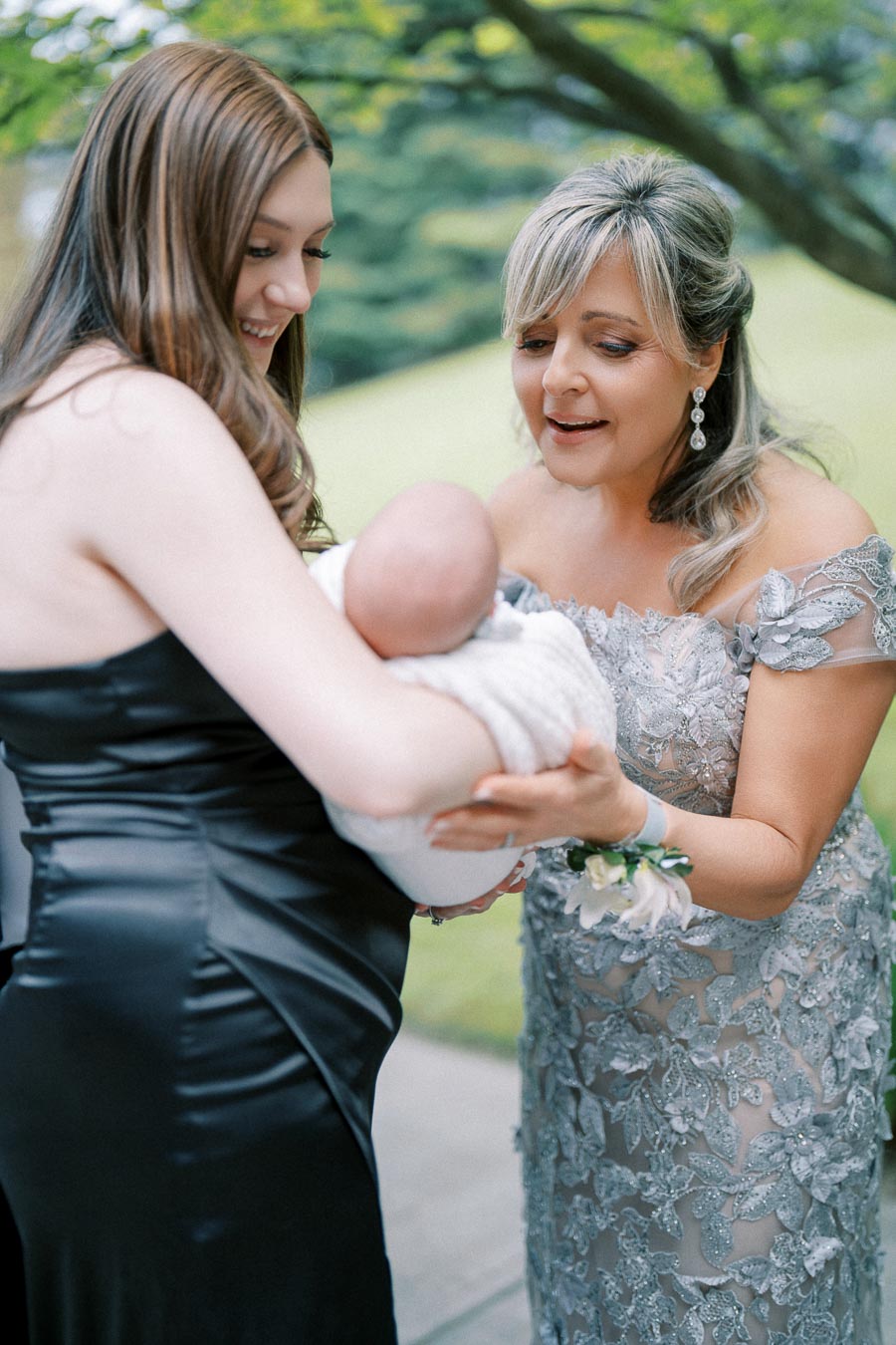 Two women, one in a black dress and the other in a silver lace gown, joyfully admiring a baby wrapped in a white blanket outdoors.