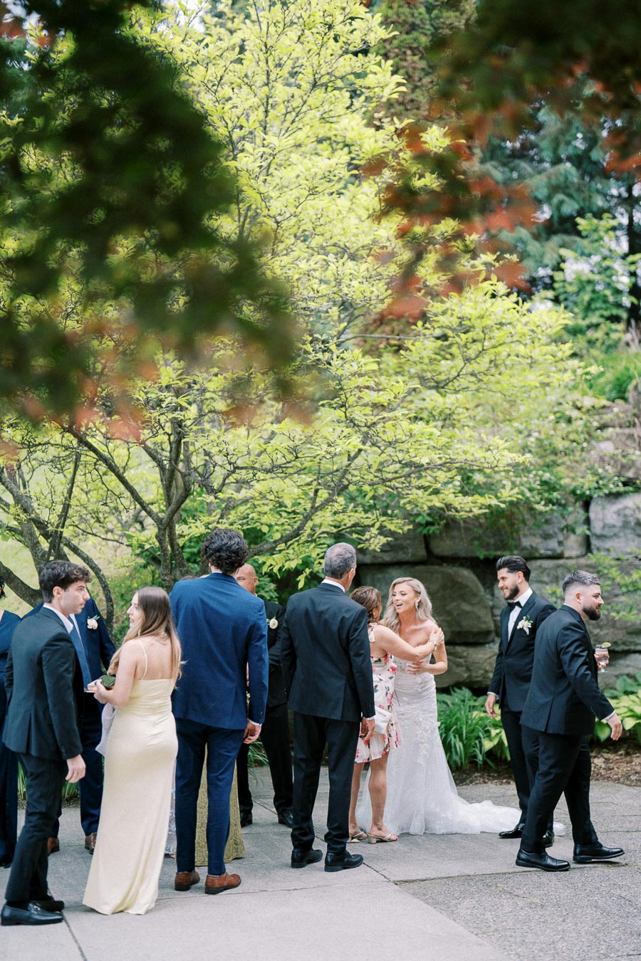 Wedding guests socializing outdoors, surrounded by lush greenery and large stone walls.