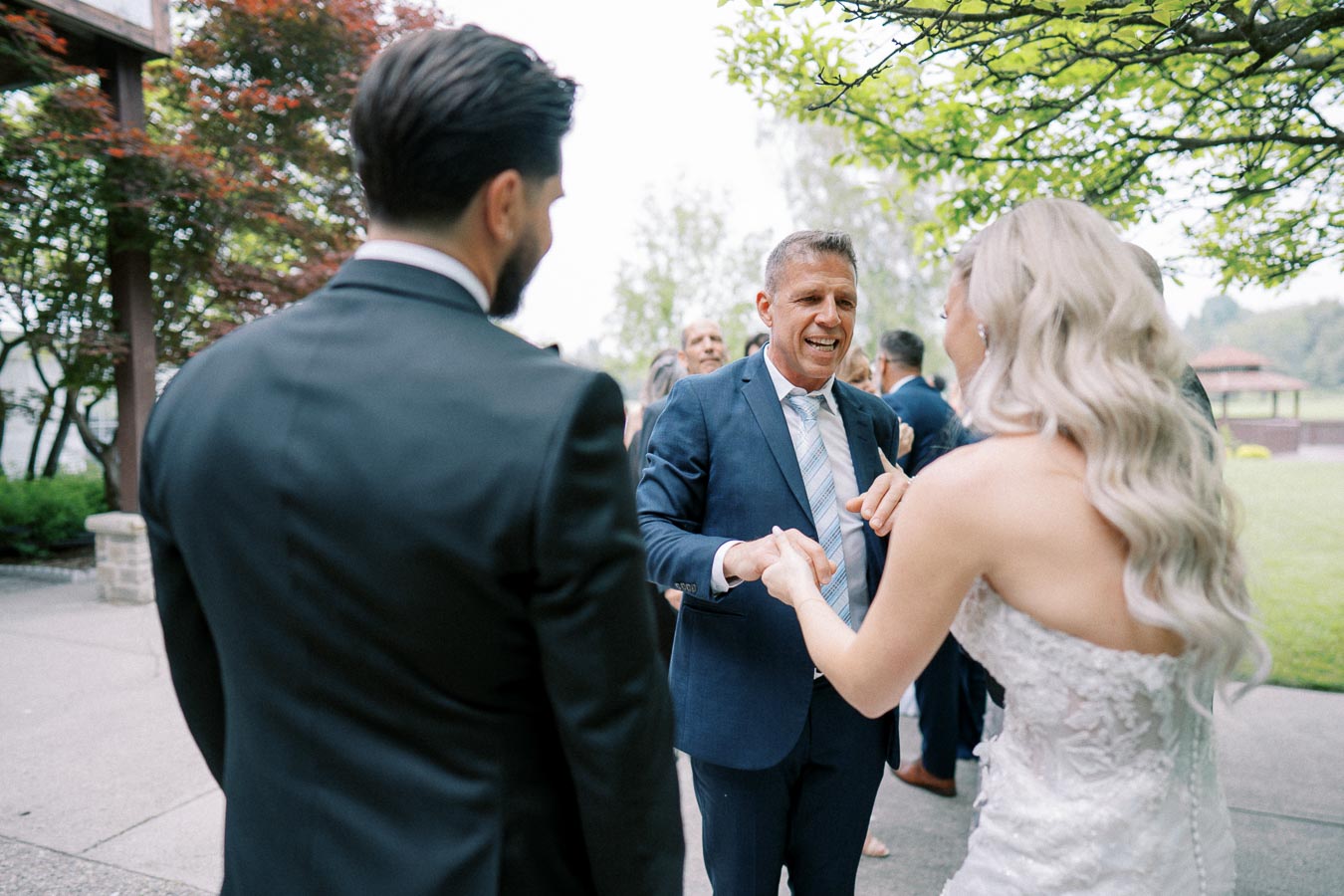 Wedding celebration scene with a bride in a white gown and long, flowing hair greeting guests outdoors. A groom in a dark suit stands nearby, while a smiling man in a blue suit engages with the couple. Lush green trees and a gazebo are visible in the background.