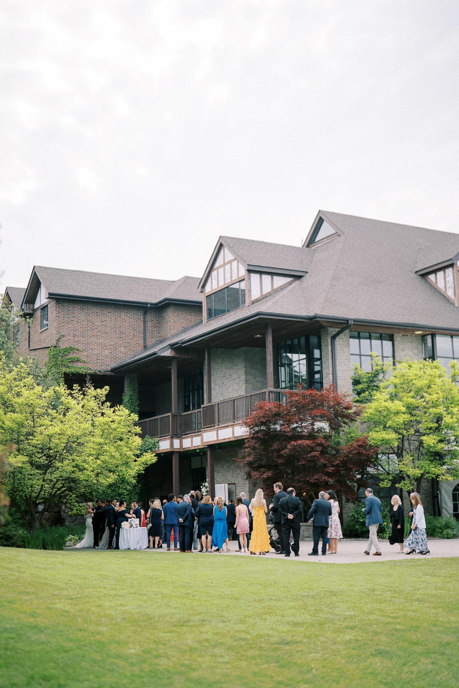 A group of elegantly dressed people gathered outside a large, classic brick building with greenery and landscaped lawns, possibly attending an outdoor event or wedding reception.