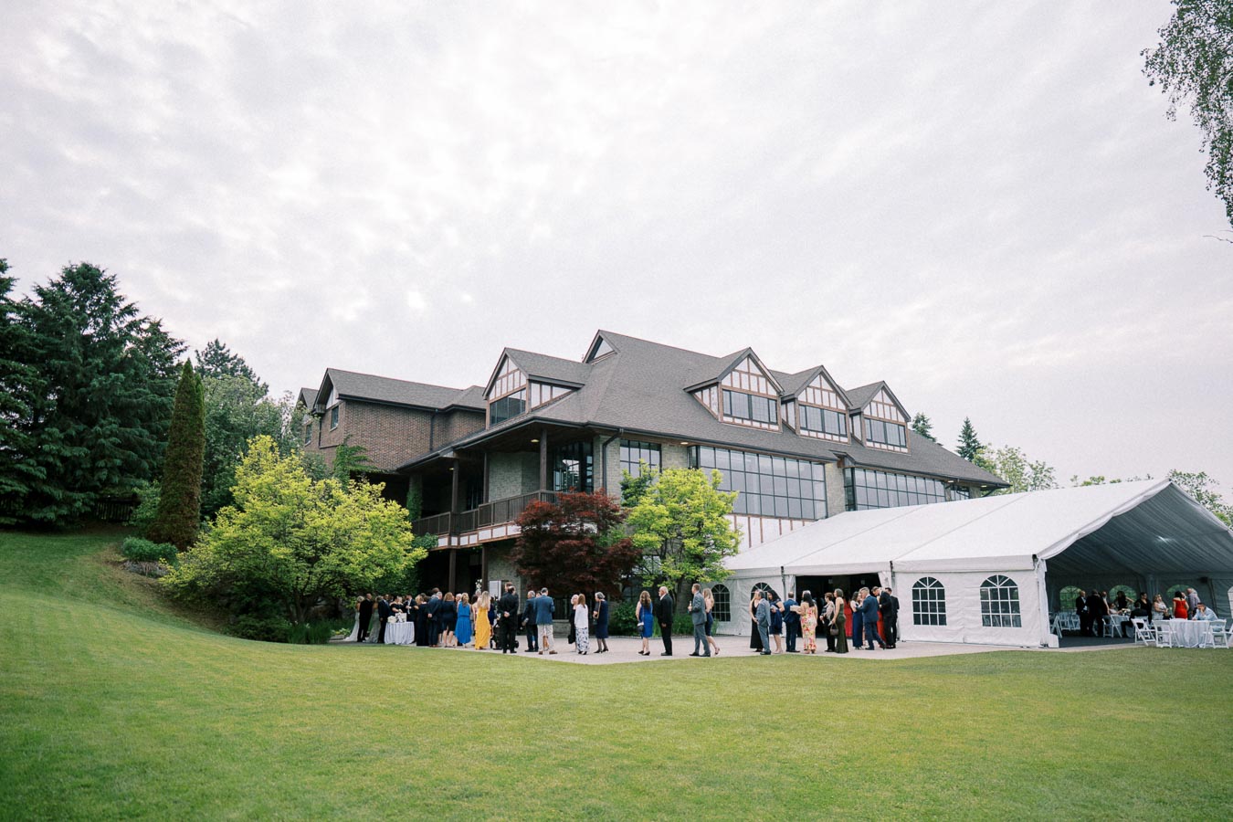 Elegant wedding reception at a picturesque estate with guests mingling outside a large white tent, surrounded by lush greenery and a grand building.