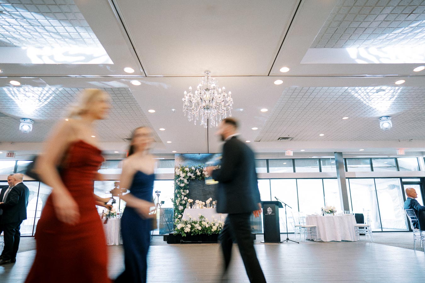 Elegant event venue with chandeliers and floral decorations, featuring guests in formal attire moving across the room.