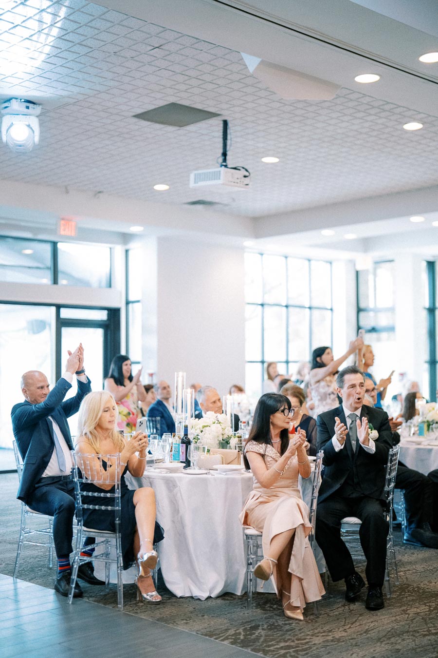 A group of elegantly dressed wedding guests clapping and taking photos at a reception, seated at a table decorated with white flowers and candles in a bright, modern venue.