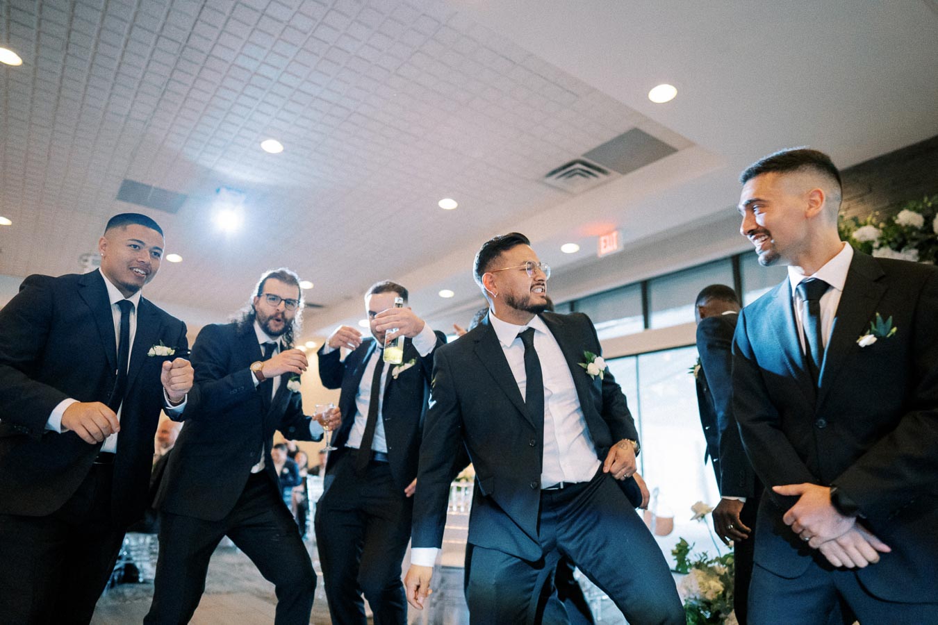A group of groomsmen in suits joyfully celebrating at a wedding reception indoors, holding drinks and smiling, with decorative flowers in the background.