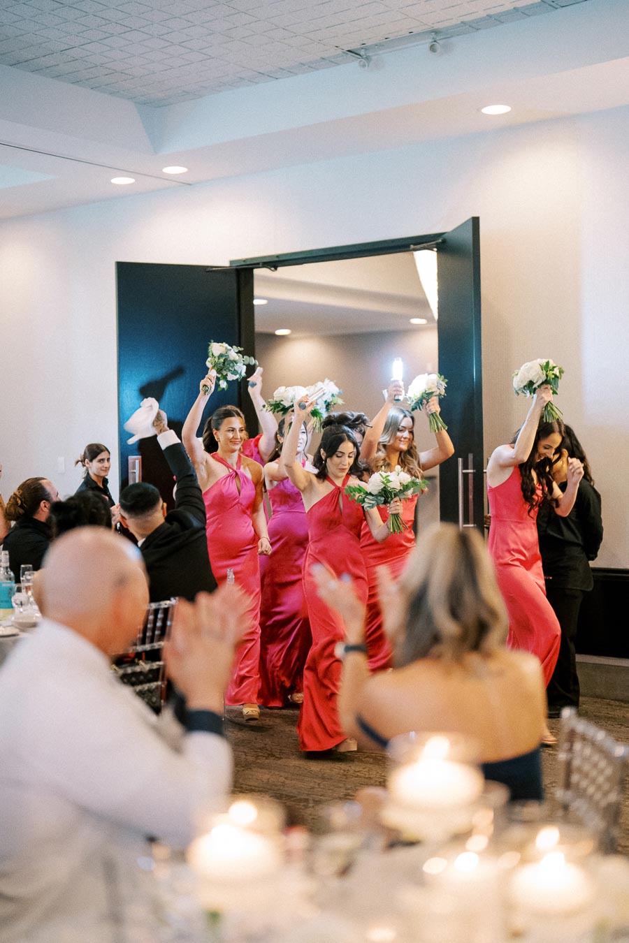 Bridesmaids in elegant pink dresses holding bouquets enter a wedding reception hall, greeted by clapping guests.