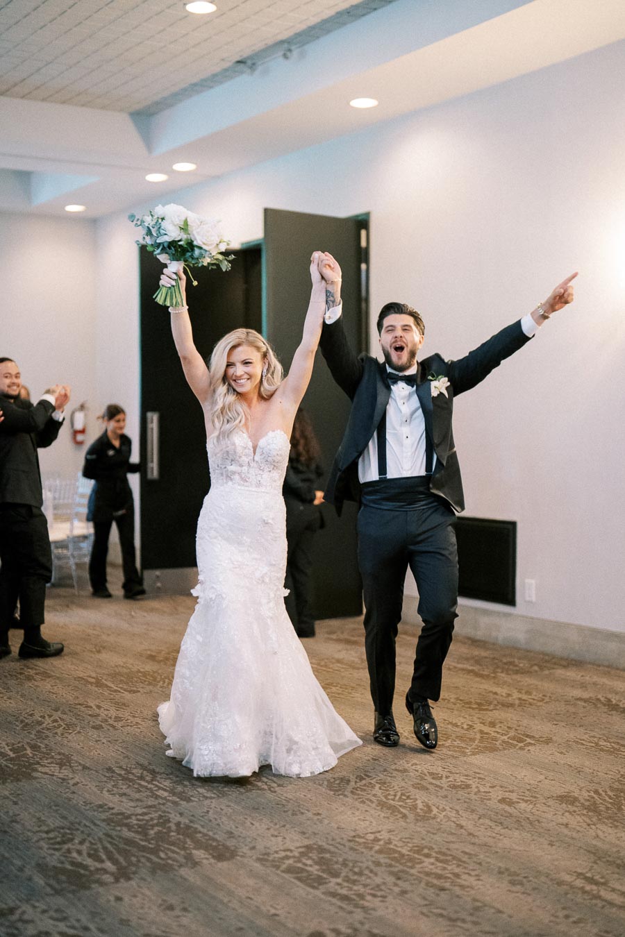Joyful bride and groom entering wedding reception with enthusiasm, bride holding bouquet, both dressed in elegant wedding attire.