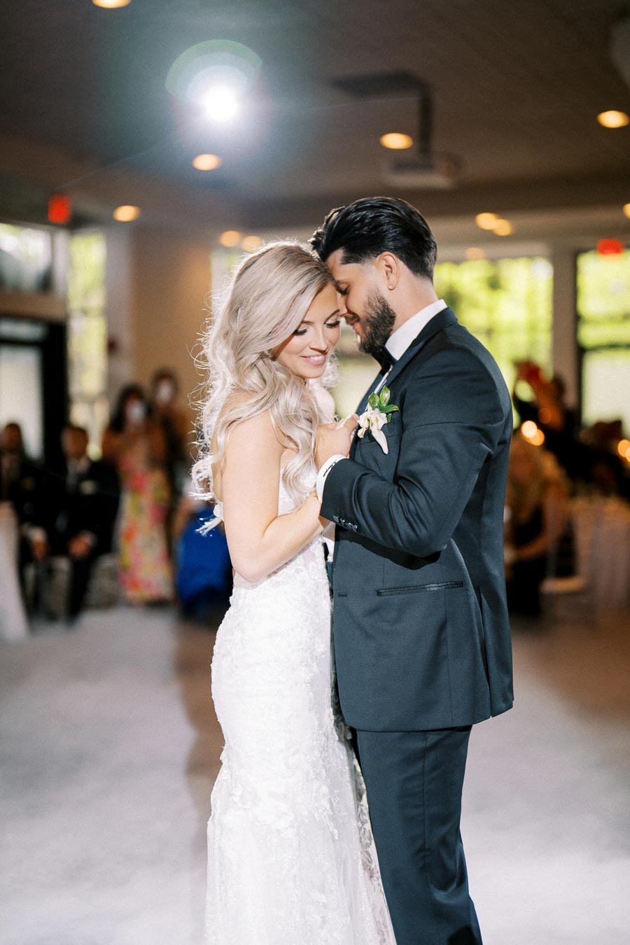 A couple sharing their first dance at a wedding reception, with the bride in a beautiful white gown and the groom in a dark suit, surrounded by warmly lit decorative lights.