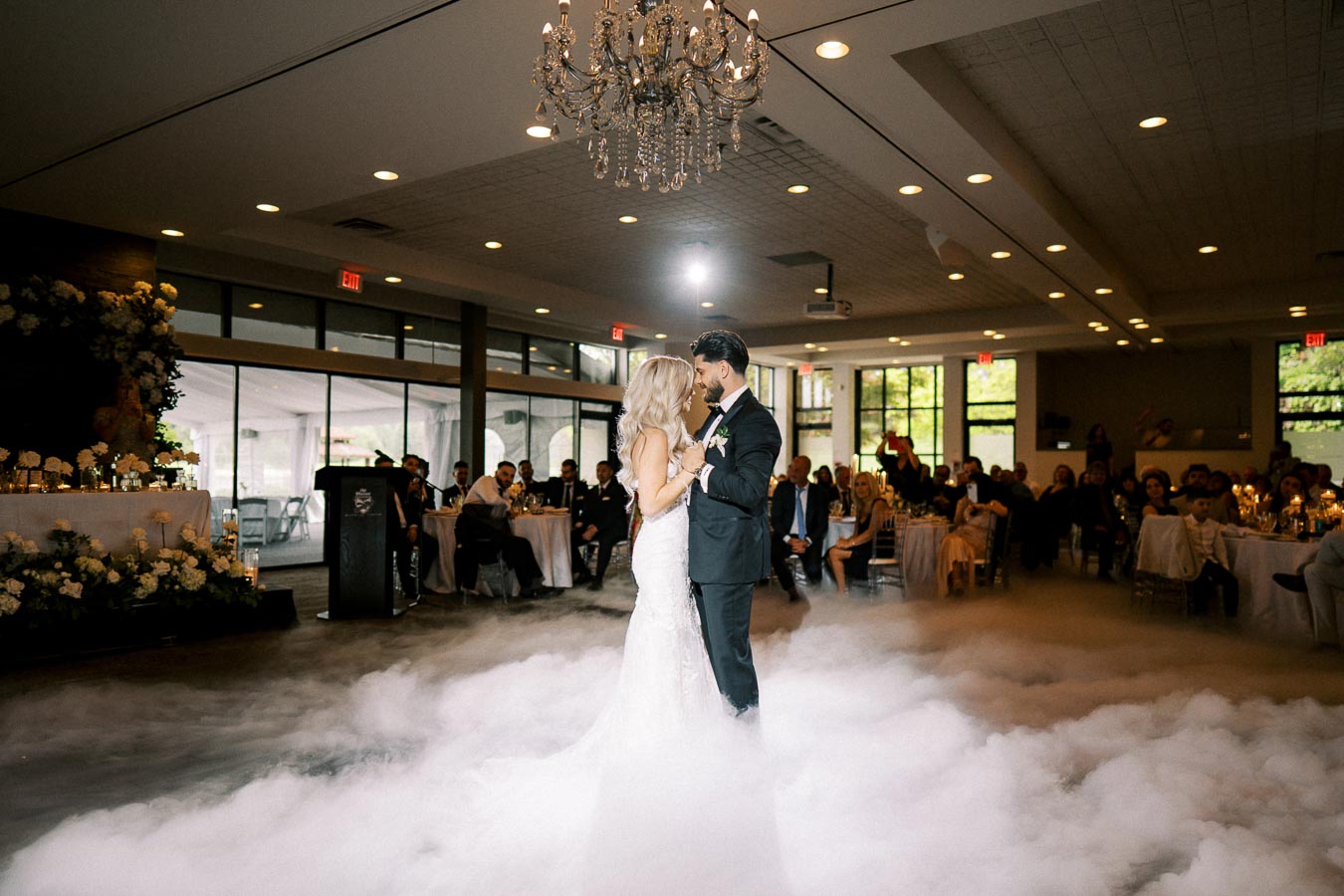 A bride and groom share their first dance surrounded by mist on the floor, under a sparkling chandelier in a beautifully decorated wedding reception venue.