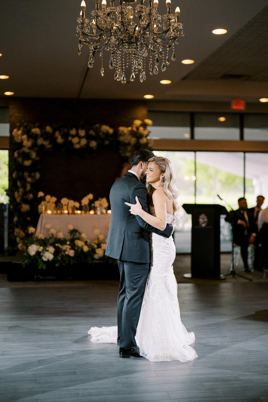 Elegant wedding couple sharing a romantic first dance under a sparkling chandelier, surrounded by floral arrangements and soft candlelight in a beautifully decorated venue.