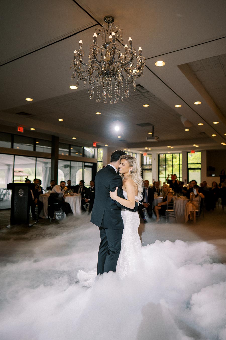 A couple shares their first dance on a cloud-like floor under a chandelier in a beautifully decorated wedding venue, surrounded by guests.