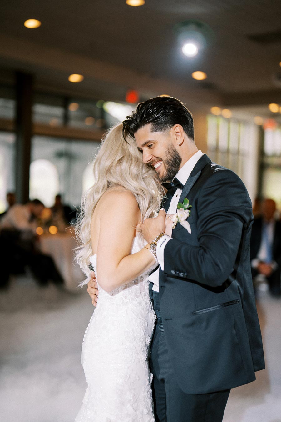 A bride and groom share a tender dance at their wedding reception, with the groom dressed in a black suit and the bride in a white lace gown, surrounded by soft lighting in a beautifully decorated venue.