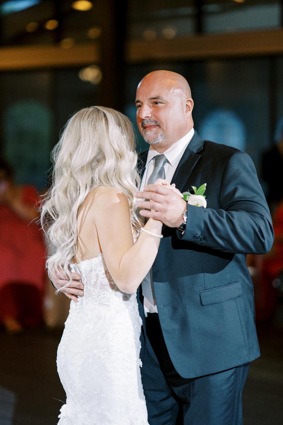 Father and bride dancing at a wedding reception, the bride wearing a white lace gown and the father in a dark suit with a boutonniere, in an elegantly lit venue.