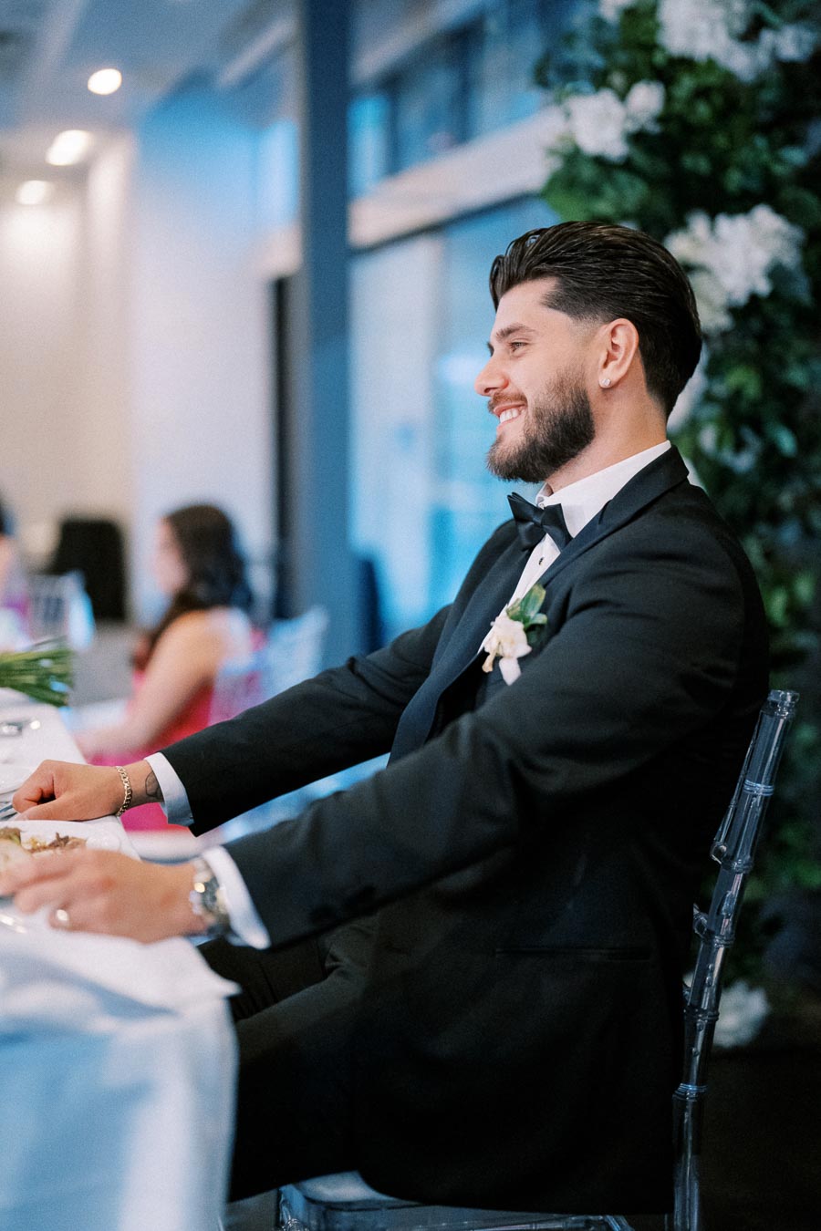 A man in a black tuxedo smiling while seated at an elegant dining table, with a floral arrangement on the wall behind him, in a formal event setting.