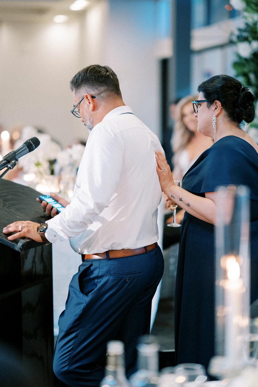 A man in a white shirt and blue pants speaks into a microphone at a podium, while a woman in a navy dress stands beside him touching his back, during an indoor formal event.