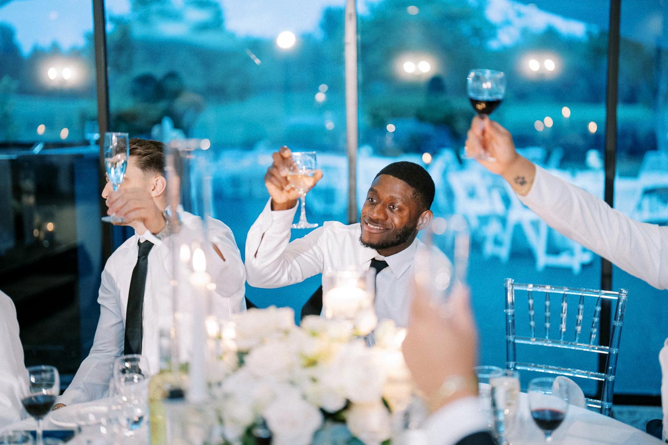 Guests in formal attire raising glasses for a toast at an elegant indoor event with a scenic outdoor backdrop.