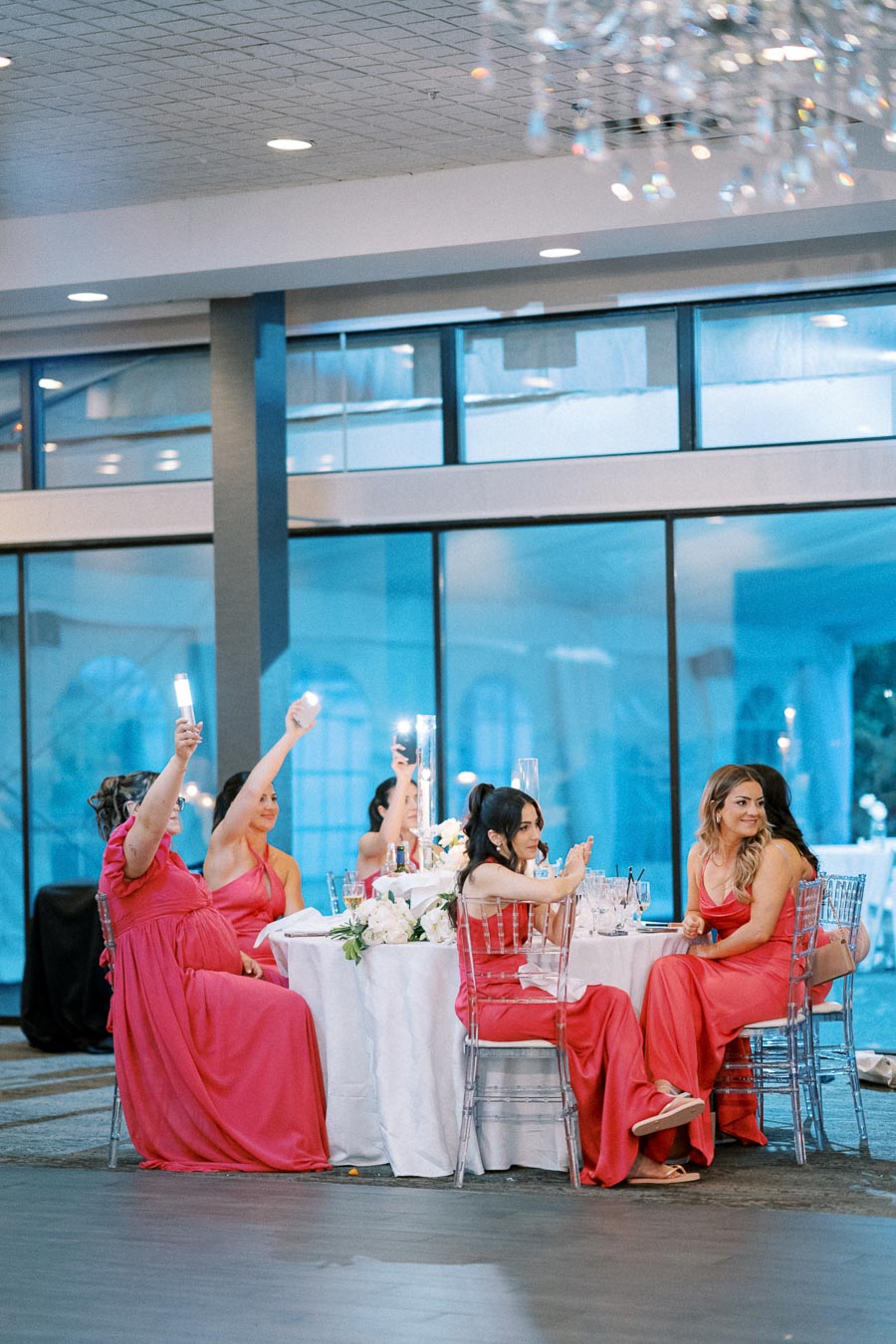 Bridesmaids in matching pink dresses sitting at a decorated reception table, raising candles in a celebratory gesture inside a modern banquet hall with large windows.