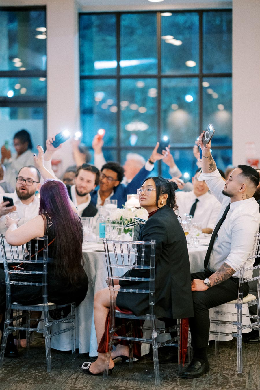 A group of people sitting at a formal event taking photos and videos with their phones, surrounded by elegant table settings and large windows in the background.