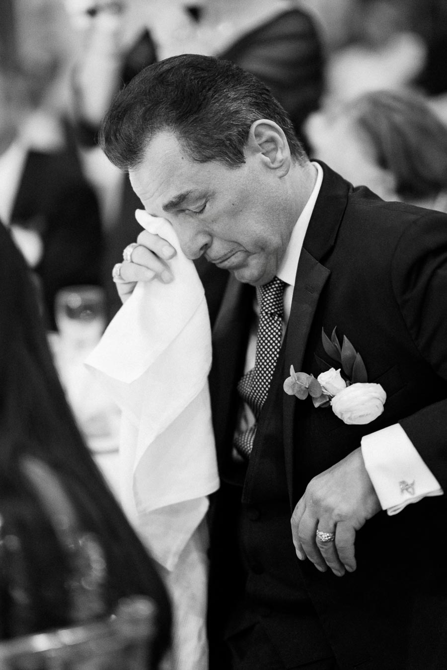 A man in a formal suit wipes tears with a napkin during an emotional moment at a wedding reception.